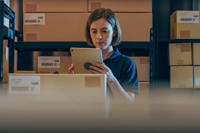 Person using a tablet in a warehouse setting with boxes and shelves.