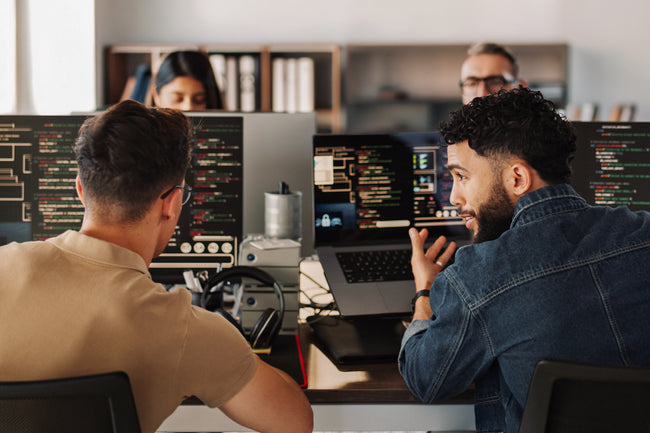 Two men working at computers in a tech support or coding office setting.